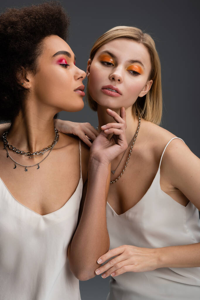 sensual african american woman in white strap dress and silver necklaces touching chin of blonde model with bright makeup isolated on grey - Photo, image