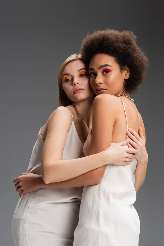 multietnické blondýny a brunetky modely v bílých kuklách a světlé tváře objímající izolované na šedé - Fotografie, Obrázek