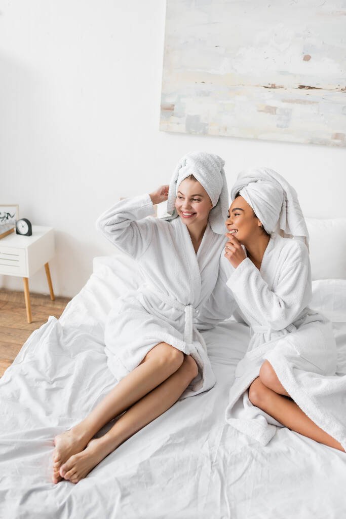 положительный африканский американец женщина в белом халате и полотенце разговаривает с улыбающимся другом глядя в сторону на кровать - Фото, изображение