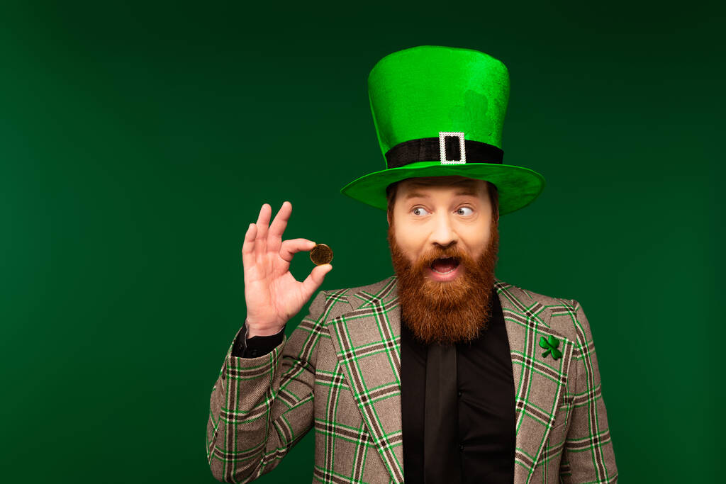 Εκπληκτικός γενειοφόρος άνδρας με καπέλο και σακάκι με τριφύλλι που κρατά το κέρμα απομονωμένο στο πράσινο  - Φωτογραφία, εικόνα