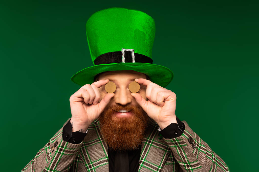 Usmívající se vousatý muž v klobouku drží mince v blízkosti očí izolovaných na zelené  - Fotografie, Obrázek