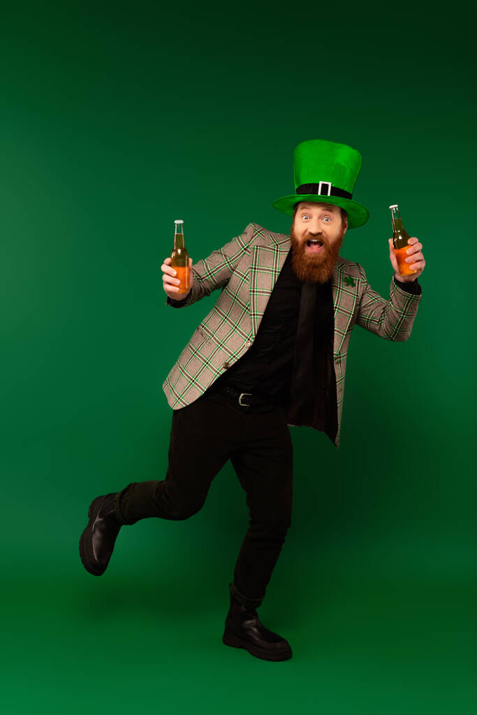 Longitud completa del hombre excitado en sombrero celebrando el día de San Patricio y sosteniendo botellas de cerveza sobre fondo verde  - Foto, imagen
