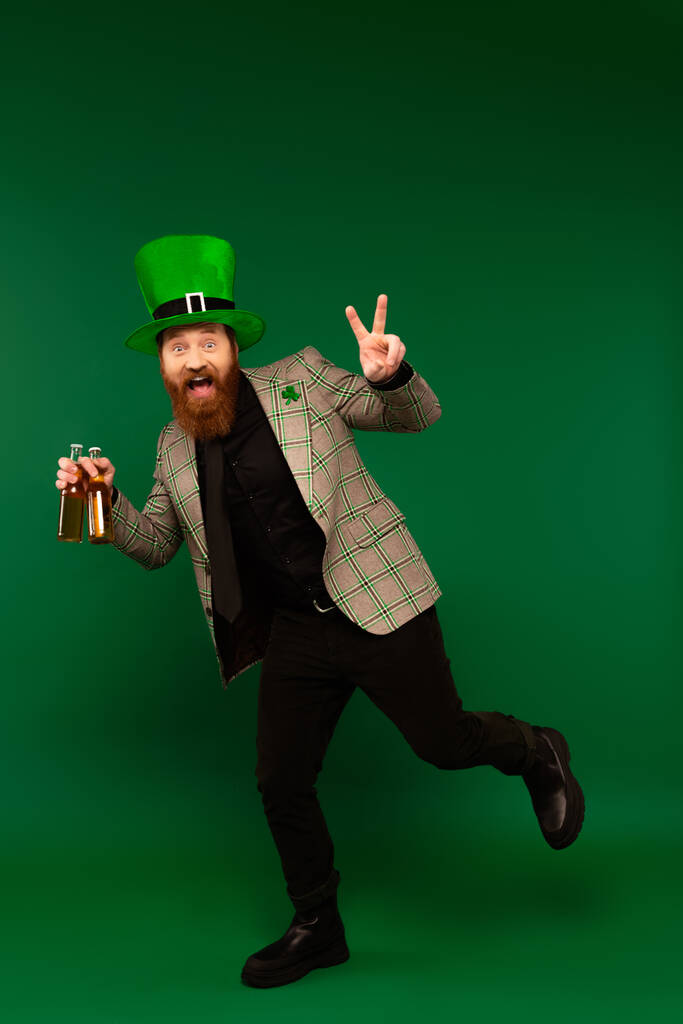 Excited bearded man in hat holding bottles of beer and showing peace sign on green background  - Fotoğraf, Görsel