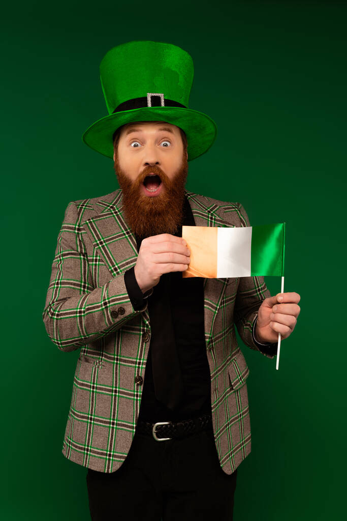 Shocked bearded man in hat holding Irish flag isolated on green  - Fotó, kép