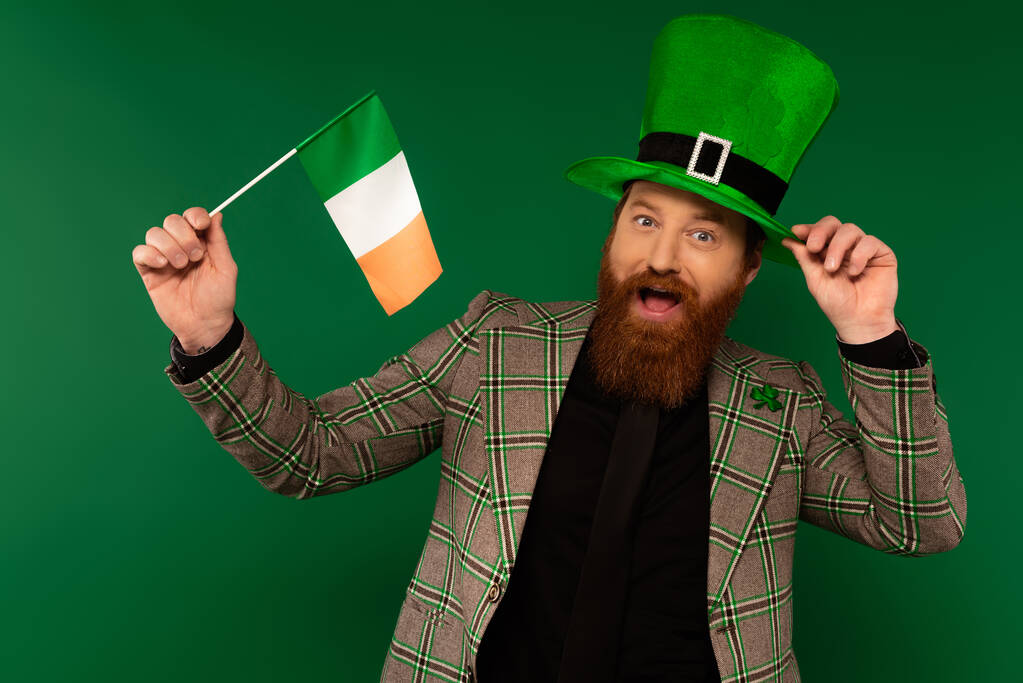 Excited bearded man in hat holding Irish flag during saint patrick day isolated on green  - Foto, imagen