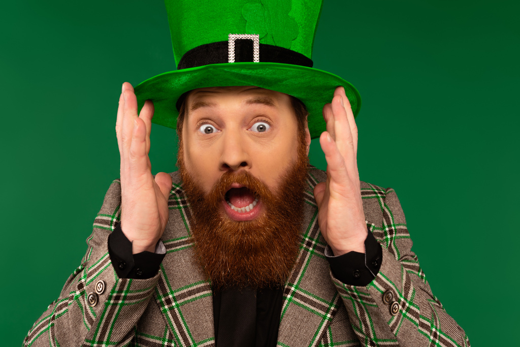 Hombre barbudo asustado en sombrero con trébol mirando a la cámara aislada en verde  - Foto, Imagen