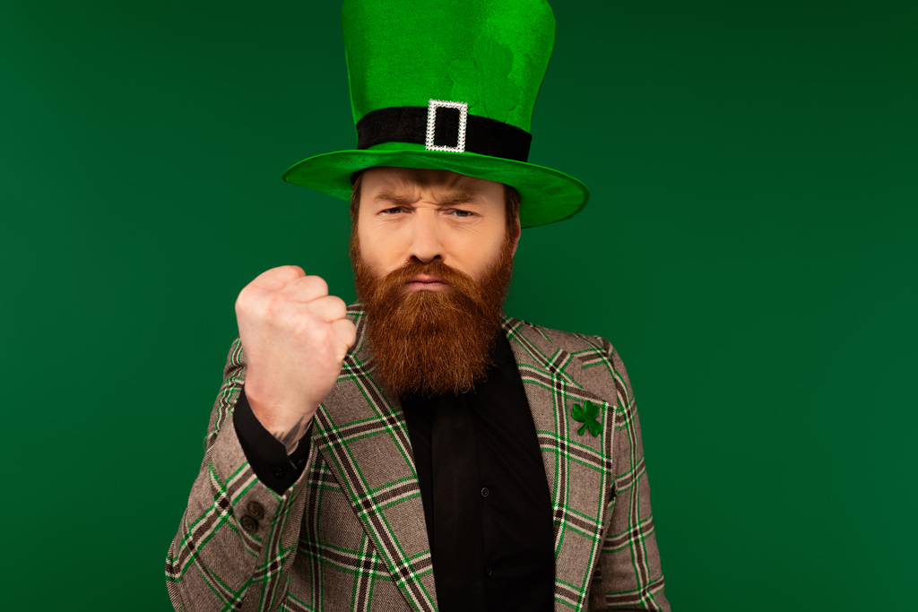 Злий бородатий чоловік в капелюсі з конюшиною, що показує кулак ізольований на зеленому
  - Фото, зображення