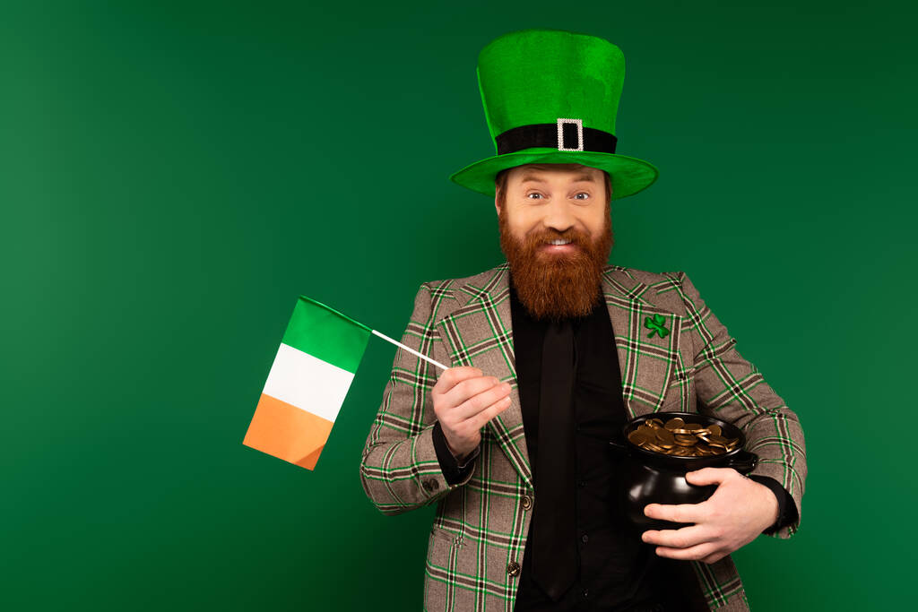 Позитивний чоловік у капелюсі з конюшиною з ірландським прапором і горщиком з монетами ізольованими на зеленому.  - Фото, зображення