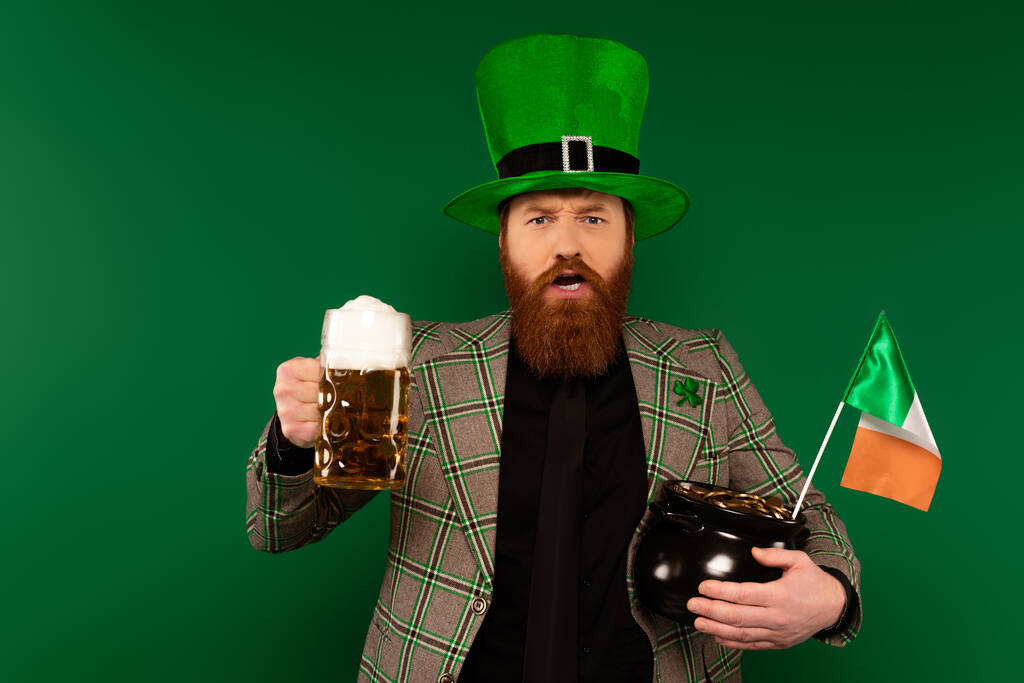 Бородач в шляпе с пивом и горшком с монетами и ирландским флагом, изолированным на зеленом  - Фото, изображение