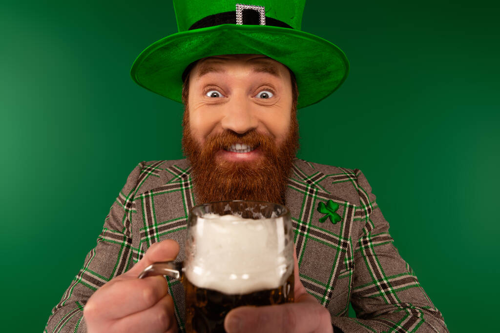 Cheerful bearded man in hat holding beer and looking at camera during saint patrick day isolated on green  - Zdjęcie, obraz