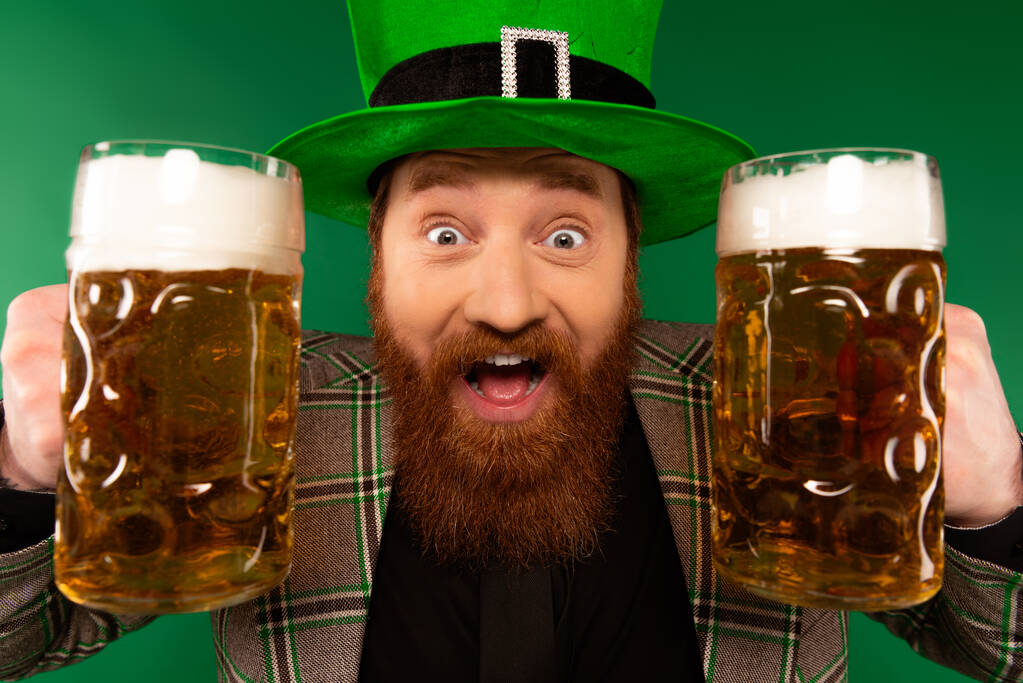 Portrait of excited man in hat holding beer glasses isolated on green  - Foto, imagen