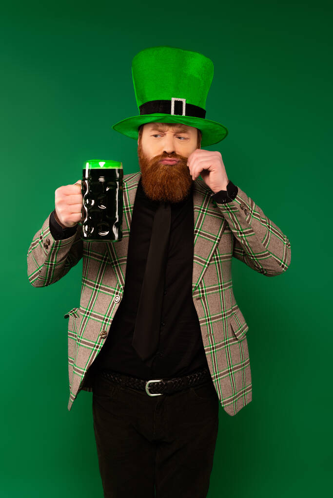 Pensive man in hat with clover looking at beer and touching moustache isolated on green  - Photo, image