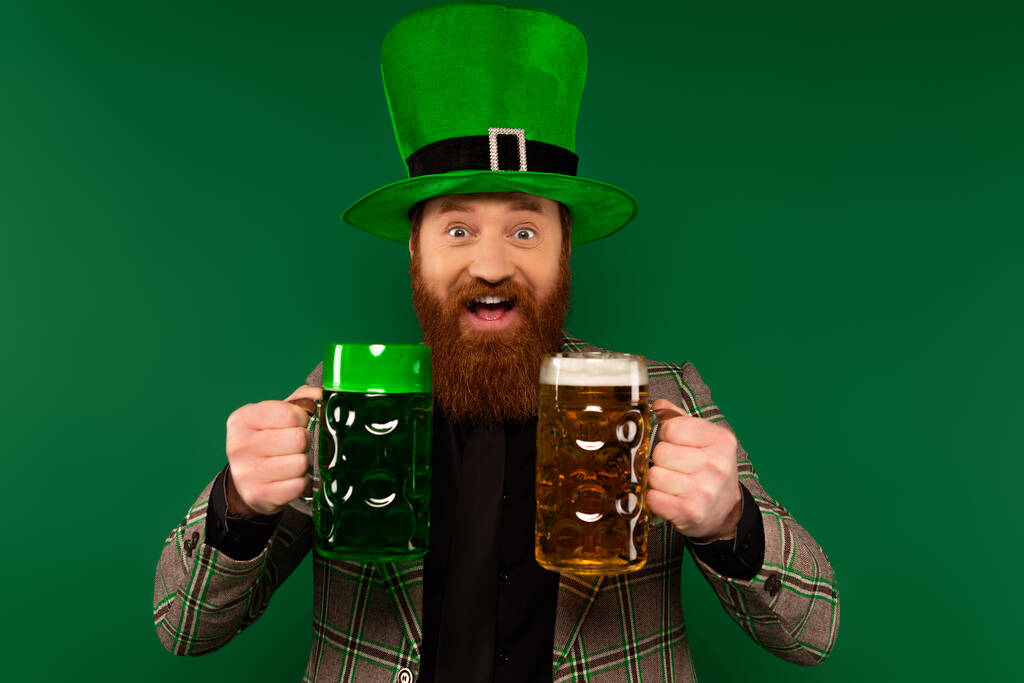 Un homme positif dans un chapeau tenant des verres de bière pendant la fête de la Saint Patrick isolé sur vert  - Photo, image