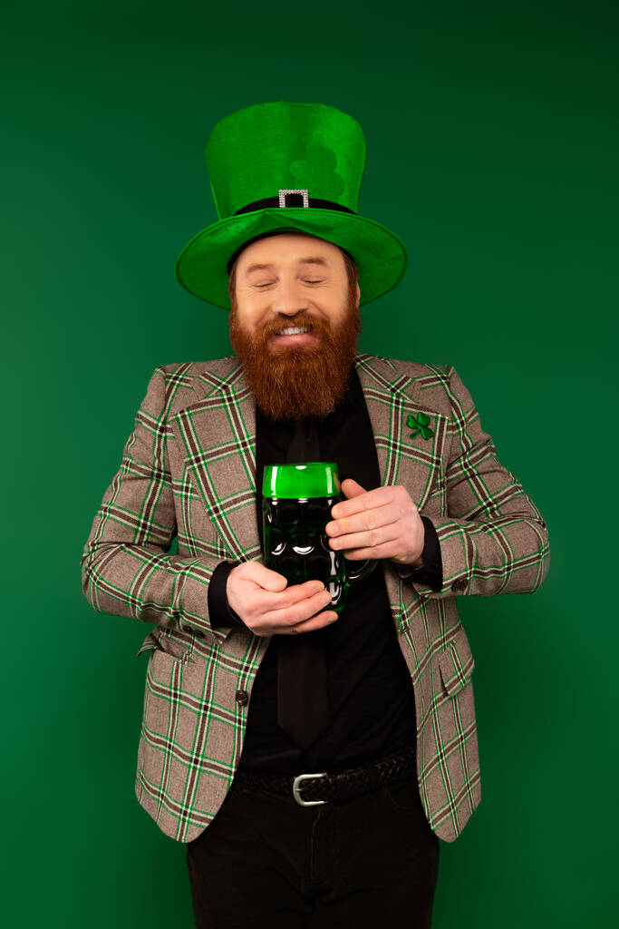 Joyful bearded man holding glass of beer while celebrating saint patrick day isolated on green  - Foto, immagini