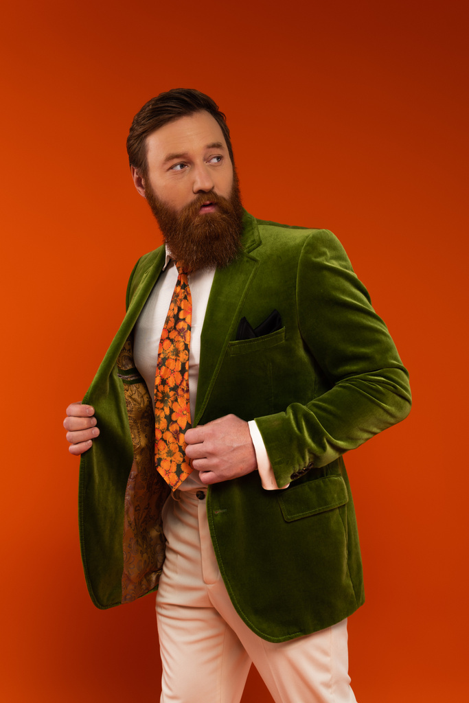 Trendy man met baard in jas weg kijkend geïsoleerd op rood  - Foto, afbeelding