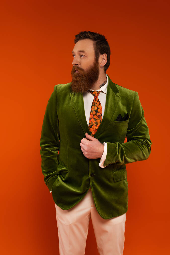 Hombre tatuado y barbudo en chaqueta verde mirando hacia otro lado aislado en rojo  - Foto, Imagen