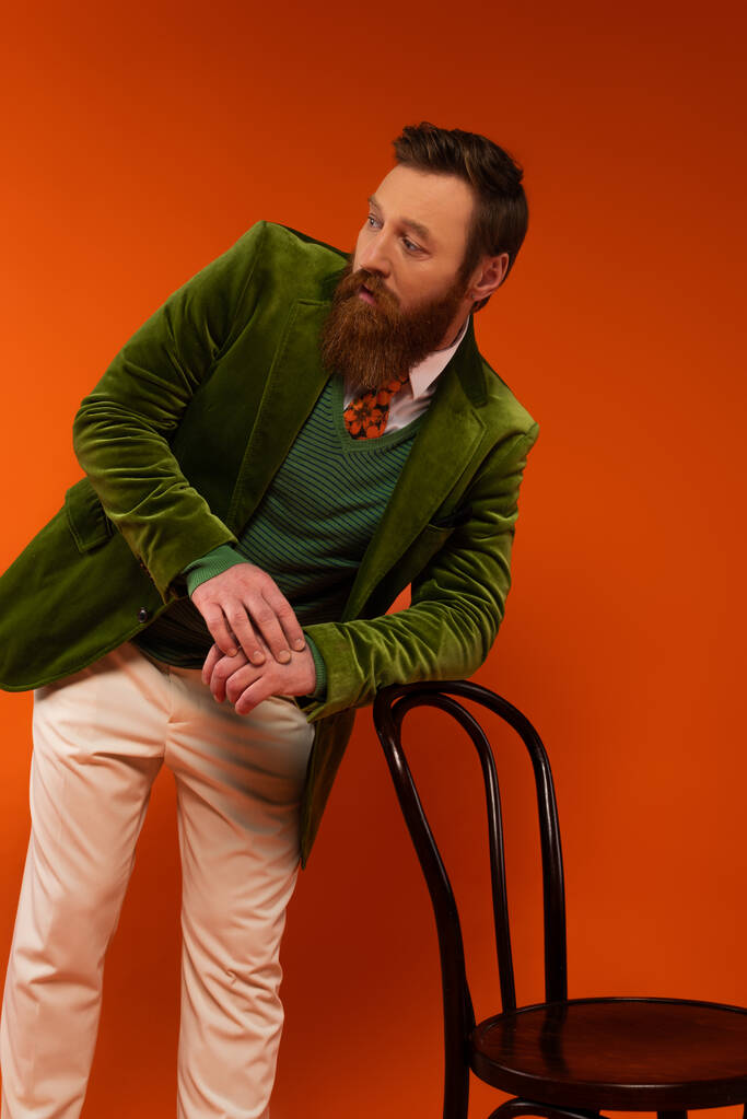 Bearded model in stylish outfit posing near chair on red background  - Fotó, kép