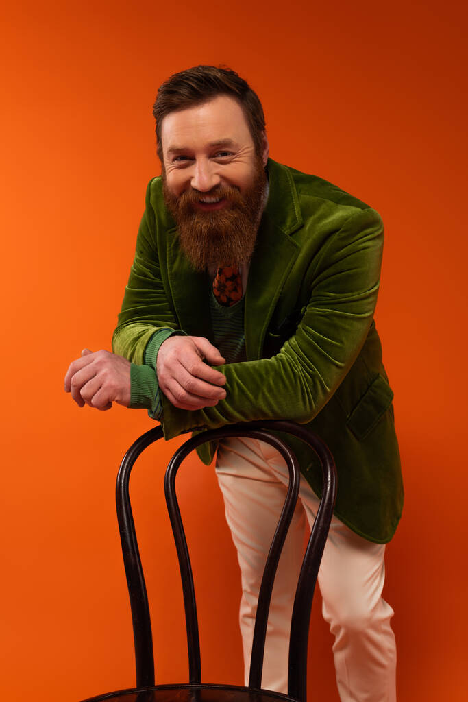 Glimlachend baard model in jas kijkend naar camera nabij stoel op rode achtergrond  - Foto, afbeelding