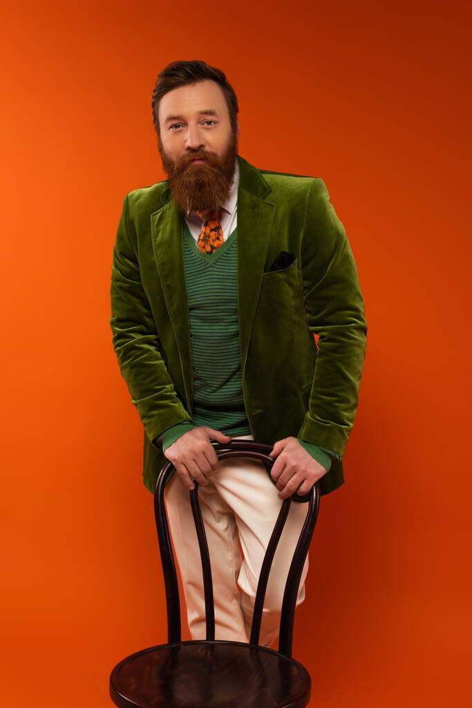Trendy bearded man standing near chair on red background  - Photo, Image