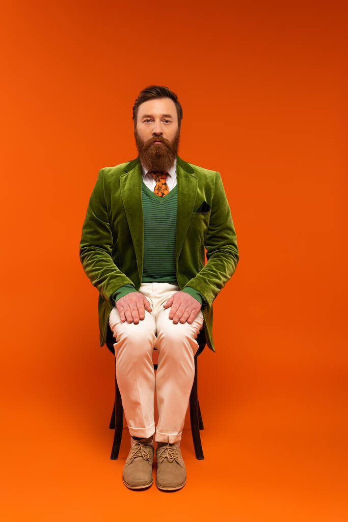 Бородатый мужчина в стильной куртке сидит на стуле на красном фоне  - Фото, изображение
