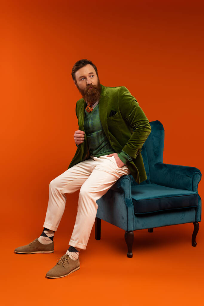 Stylish bearded man posing near velvet armchair on red background  - Fotó, kép