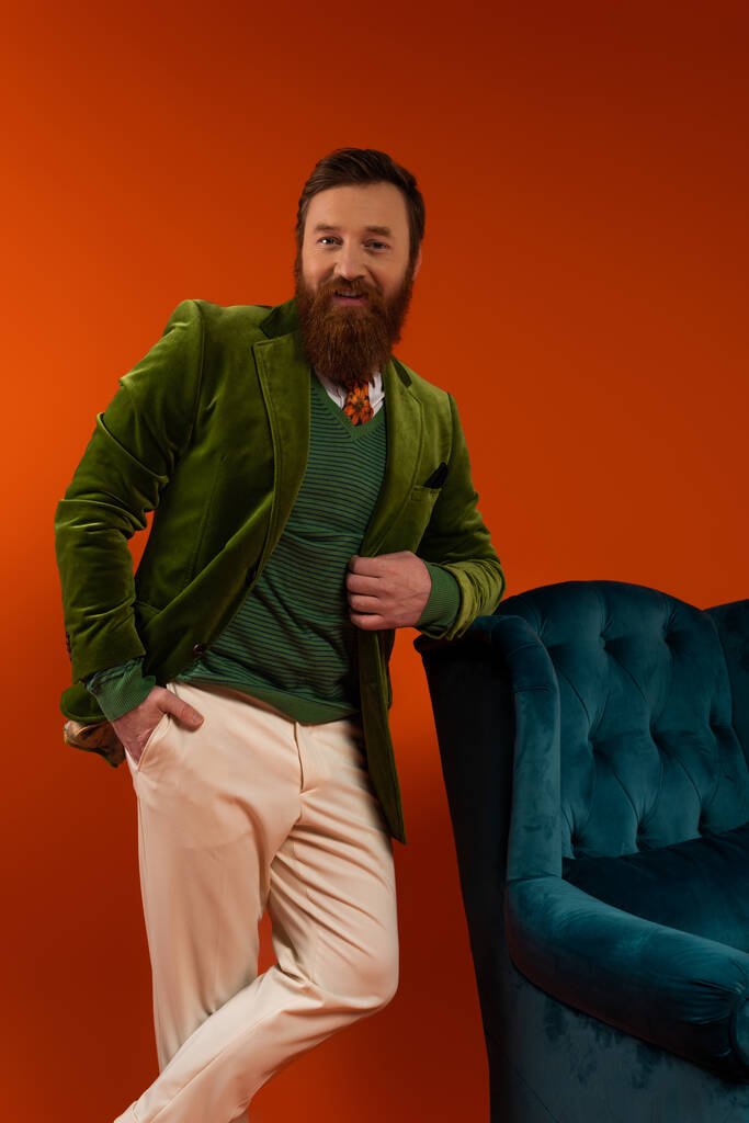 Stylish bearded man looking at camera while posing near armchair on red background  - Fotografie, Obrázek