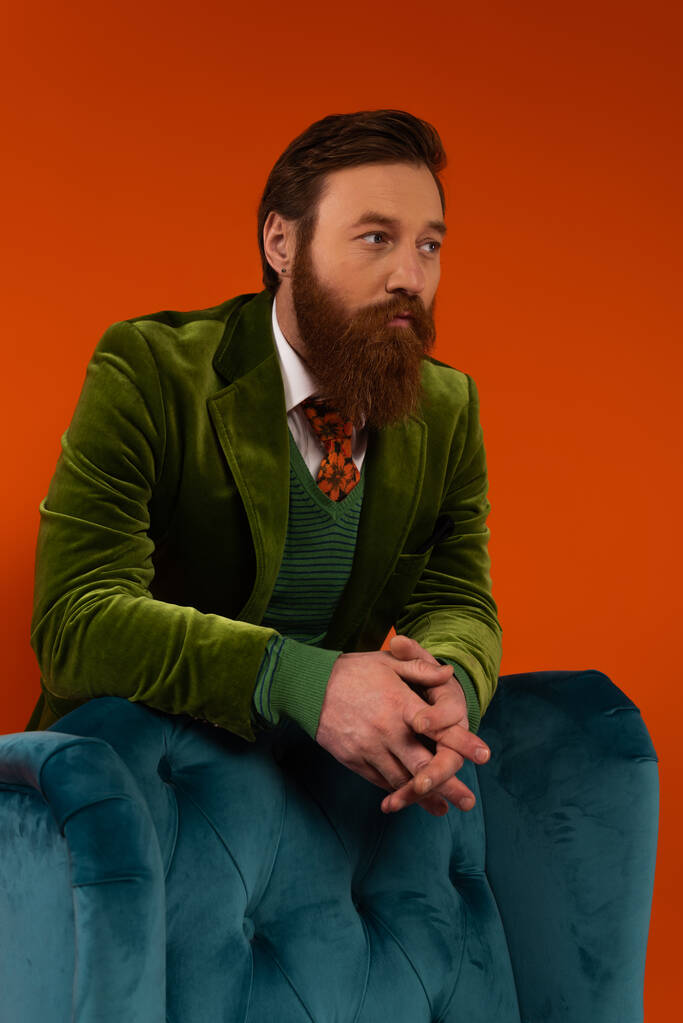 Trendy bearded model in velvet blazer standing near armchair on red background  - Foto, imagen