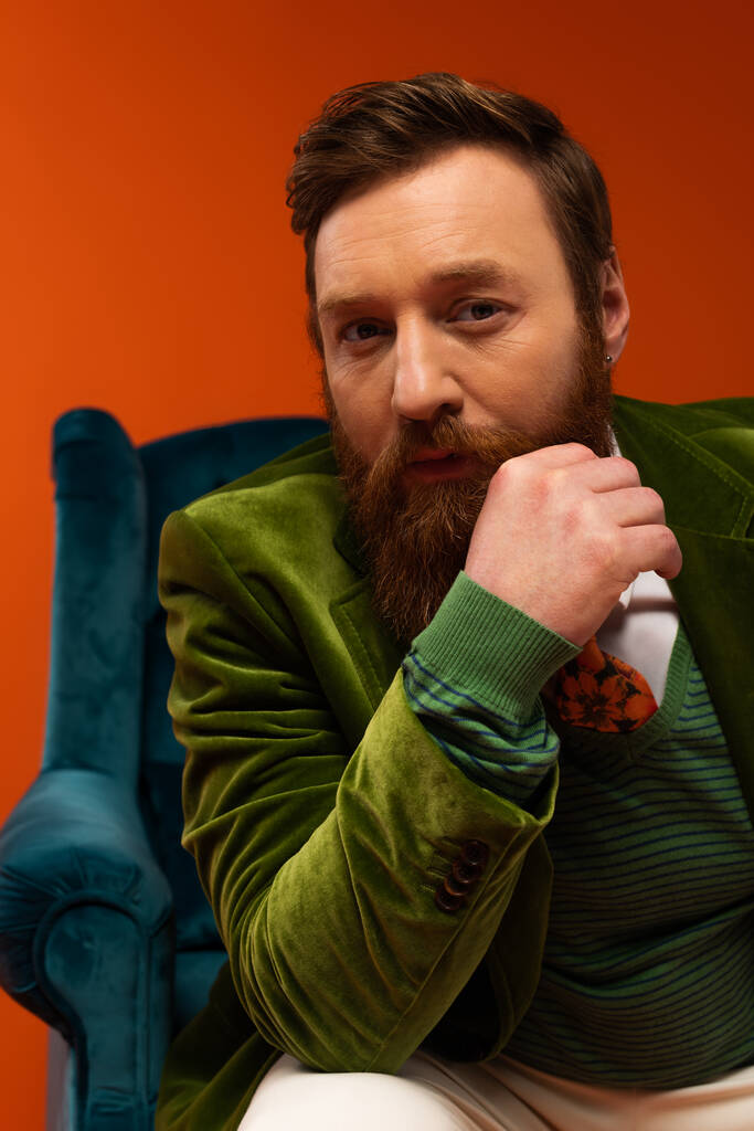 Portrait of stylish bearded model looking at camera near blurred armchair on red background  - Foto, imagen