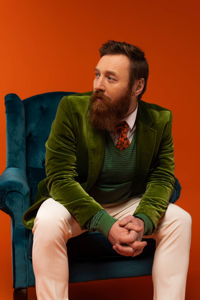 Fashionable bearded man sitting on blue velvet armchair on red background  - Foto, immagini