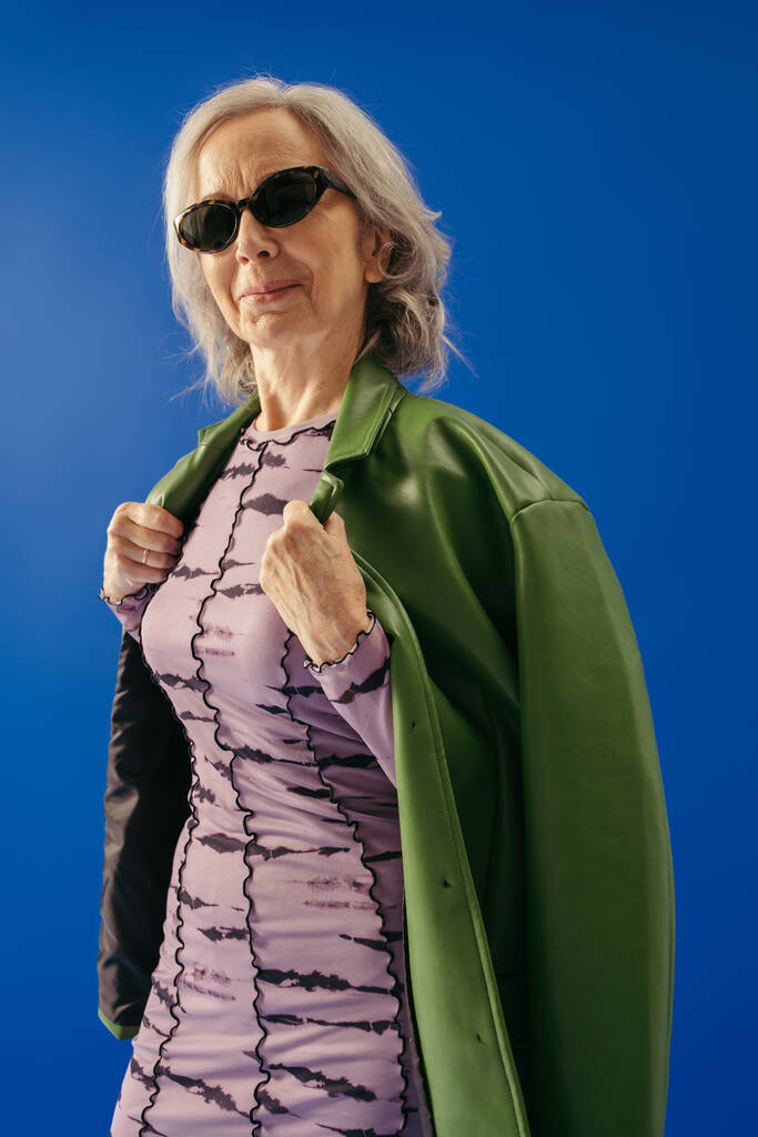 довольная пожилая женщина в солнечных очках в зеленой кожаной куртке изолированы на синий  - Фото, изображение