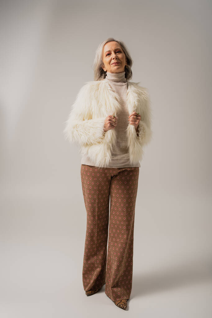 täyspitkä tyytyväinen vanhempi nainen faux turkis takki poseeraa harmaalla taustalla   - Valokuva, kuva