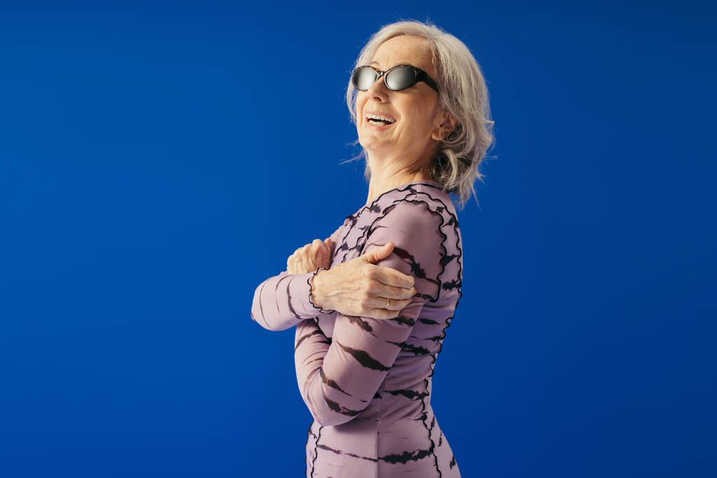 positive senior woman in sunglasses and dress looking at camera isolated on blue  - Foto, Imagem