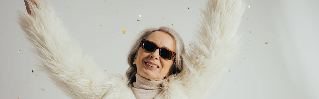 cheerful elderly woman in white faux fur jacket and sunglasses raising hands near falling confetti on grey background, banner  - 写真・画像