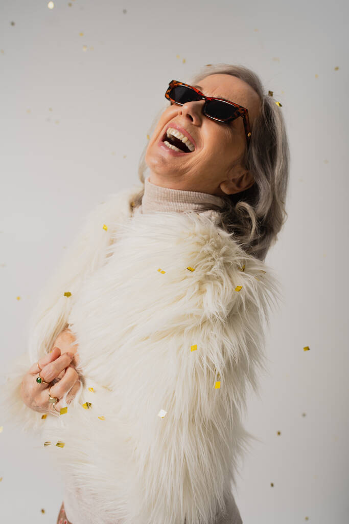 happy elderly woman in white faux fur jacket and trendy sunglasses laughing near falling confetti on grey  - Foto, Imagem