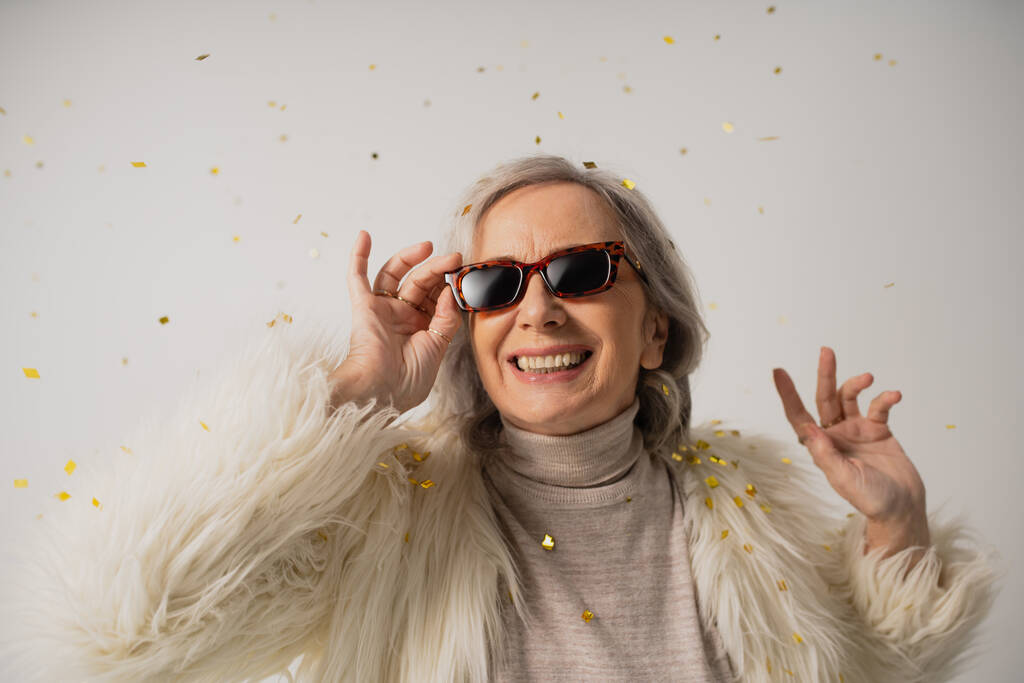 alegre anciana en chaqueta de piel sintética blanca ajustando gafas de sol de moda cerca de confeti caída sobre fondo gris  - Foto, Imagen