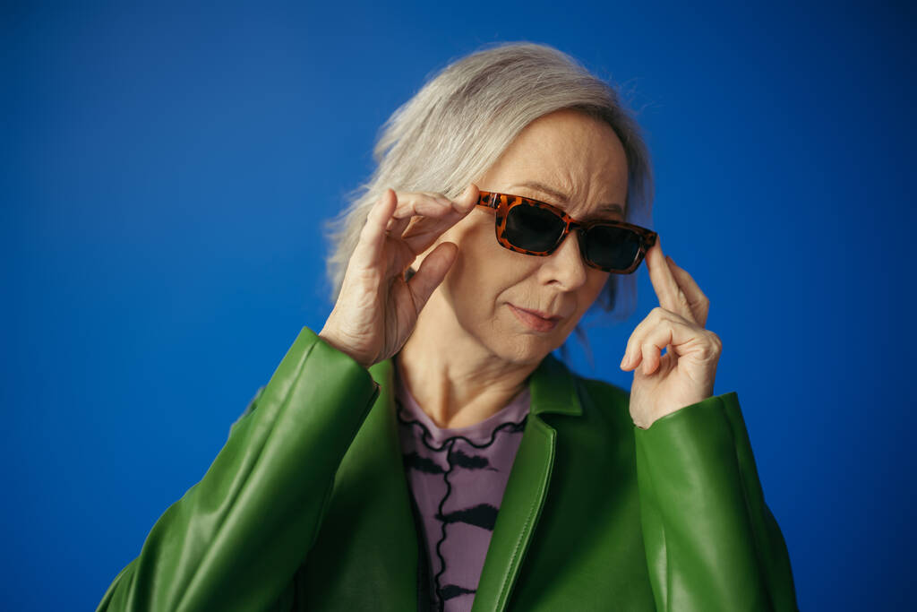 ηλικιωμένη γυναίκα με πράσινο μπουφάν που προσαρμόζει μοντέρνα γυαλιά ηλίου απομονωμένη σε μπλε - Φωτογραφία, εικόνα