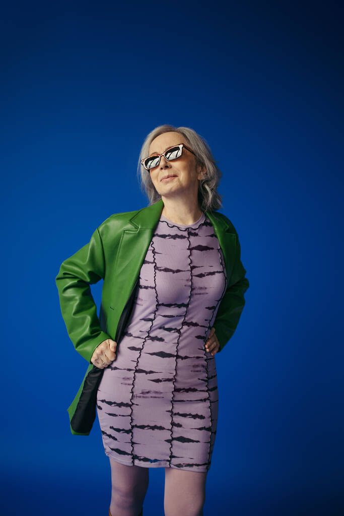 señora mayor positiva en gafas de sol de moda y chaqueta de cuero verde sobre vestido púrpura de pie con las manos en las caderas aisladas en azul - Foto, Imagen
