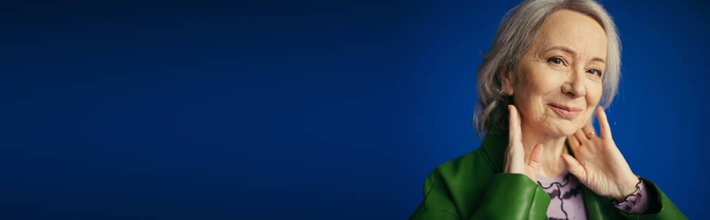 青い旗の上に孤立した首の近くに手でポーズをとる緑の革のジャケットに笑顔のシニア女性の肖像画 - 写真・画像