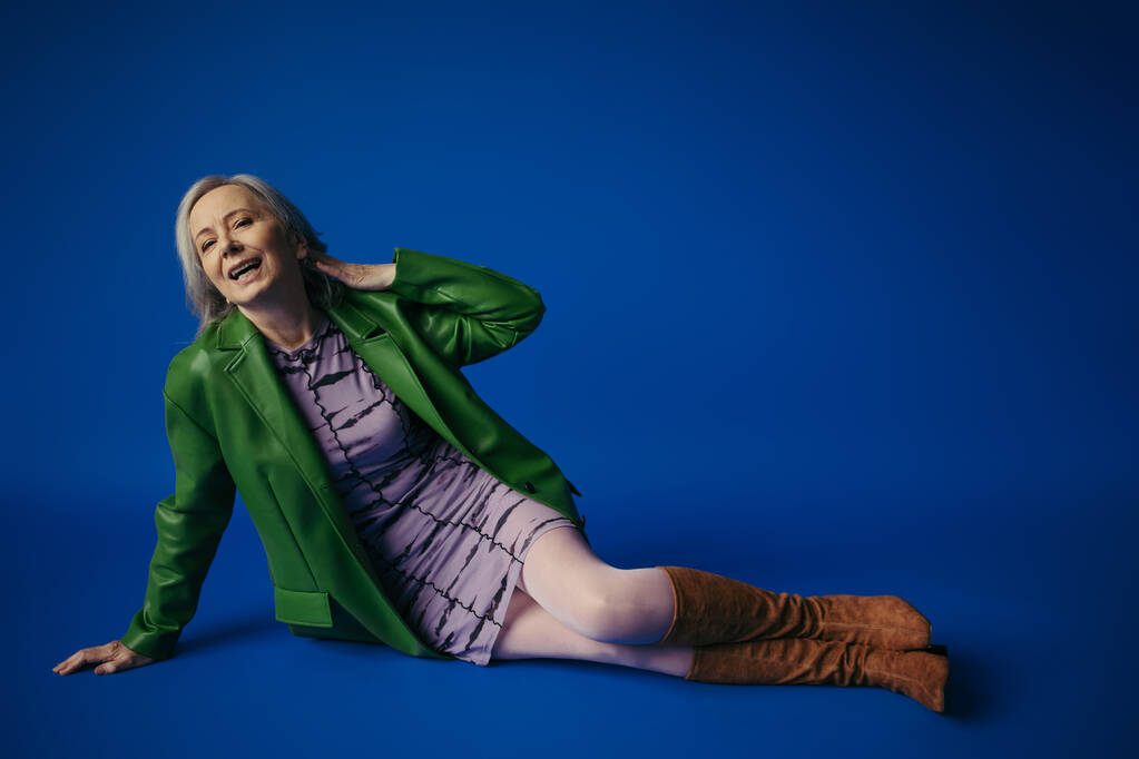 full length of cheerful senior lady in green leather jacket and purple dress sitting on blue background - Fotó, kép
