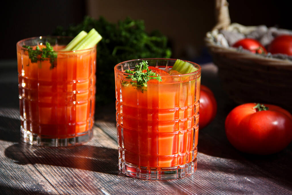 Tomatensaft in Nahaufnahme mit frischen Tomaten und Basilikum auf dunklem Hintergrund. Zwei Gläser blutiger Marzipan-Cocktail mit frischem Sellerie und Petersilie auf einem Holztisch im rustikalen Stil. Frischer Gemüsesaft mit Tomaten - Foto, Bild