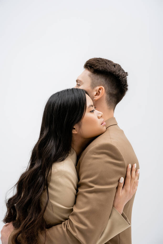 side view of brunette asian woman with long hair embracing stylish man isolated on grey - Fotó, kép