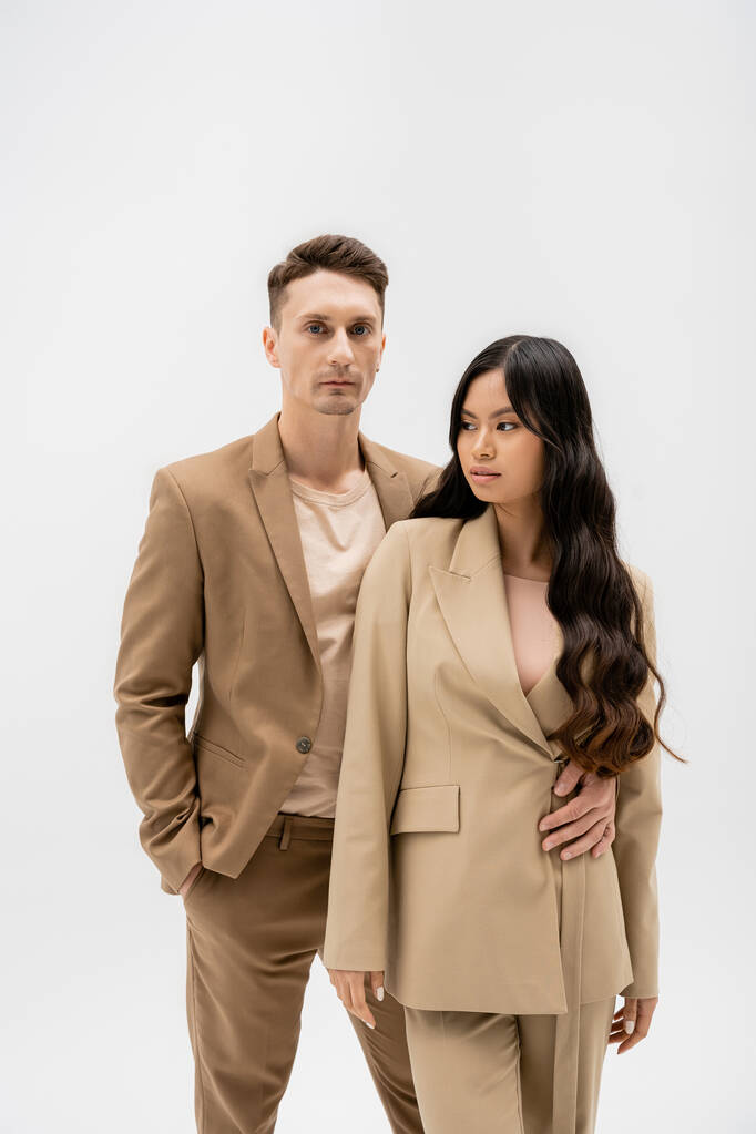 man in beige suit standing with hand in pocket and embracing asian woman isolated on grey - Fotó, kép