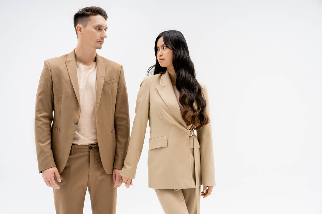 fashionable multiethnic couple in beige suits looking at each other isolated on grey - Foto, afbeelding
