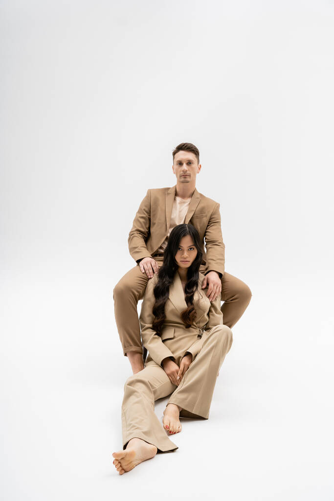 interracial couple in fashionable pantsuits looking at camera while posing on grey background - Foto, immagini