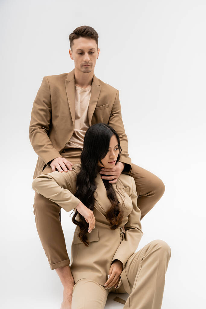 trendy man in beige suit hugging neck of young asian woman sitting on grey background - Foto, Imagem
