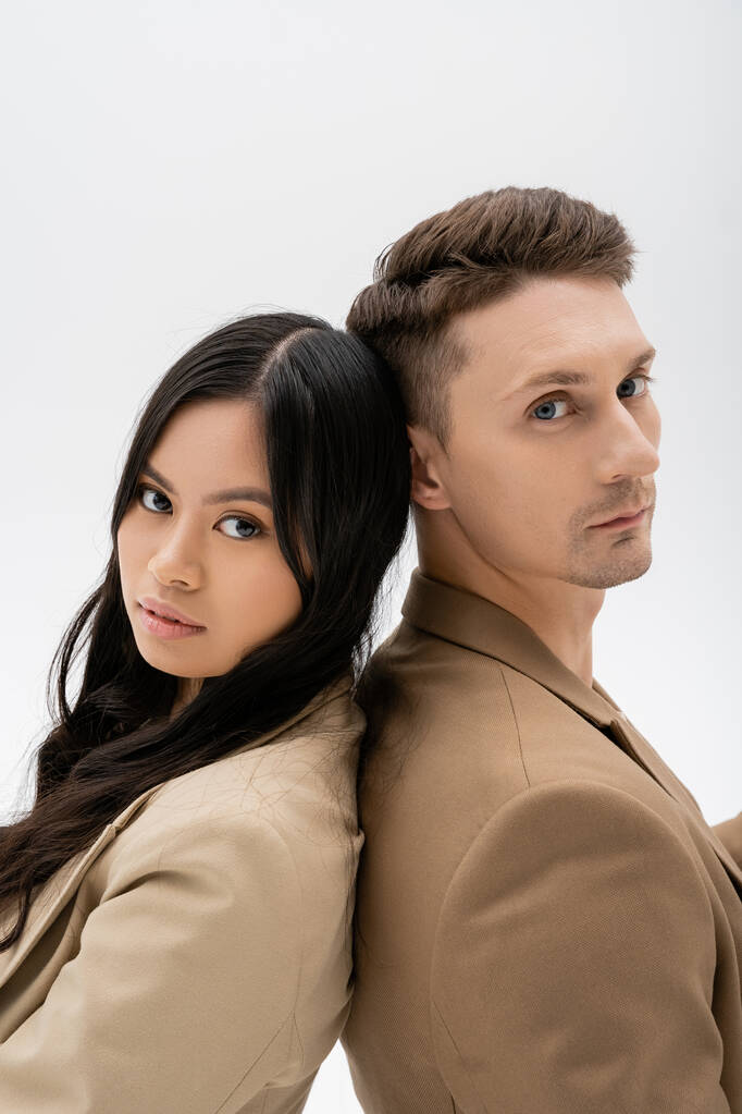 trendy interracial couple in beige jackets looking at camera isolated on grey - Fotografie, Obrázek