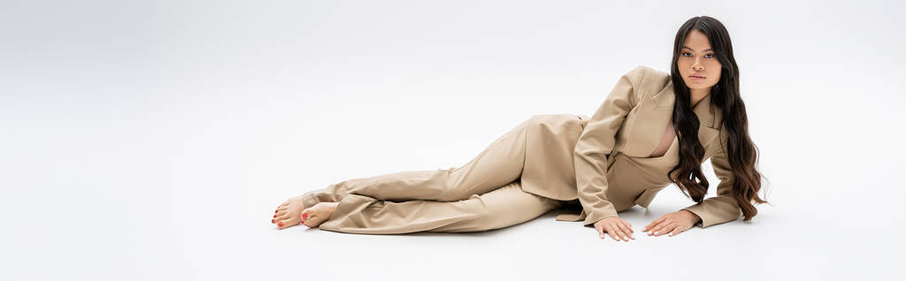 full length of brunette asian woman with long hair lying in stylish suit on grey background, banner - Foto, Bild