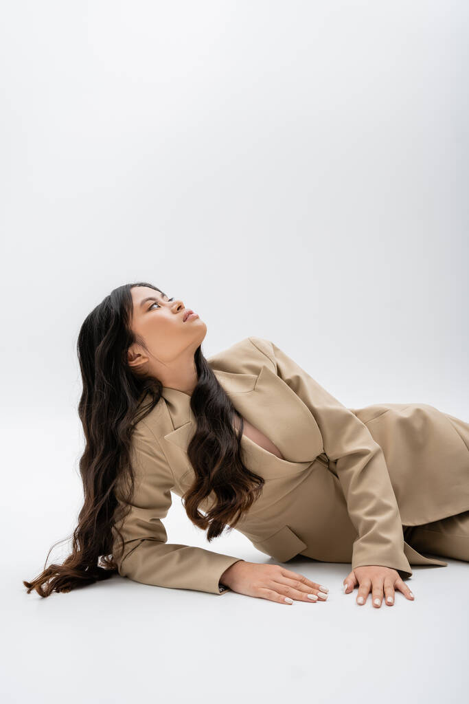 brunette asian woman in beige jacket lying and looking up on grey background - Photo, Image