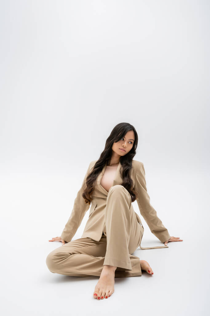 полная длина босиком азиатской женщины в бежевом костюме сидя и глядя в сторону на сером фоне - Фото, изображение