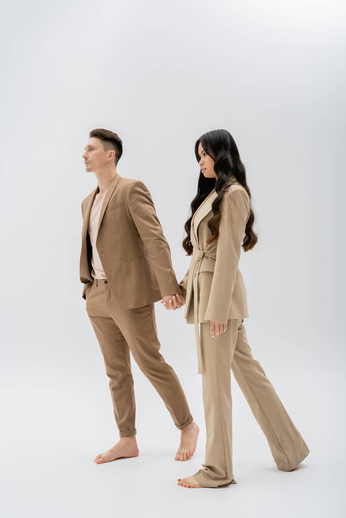 full length of barefoot multiethnic couple in beige pantsuits holding hands while walking on grey background - Foto, afbeelding